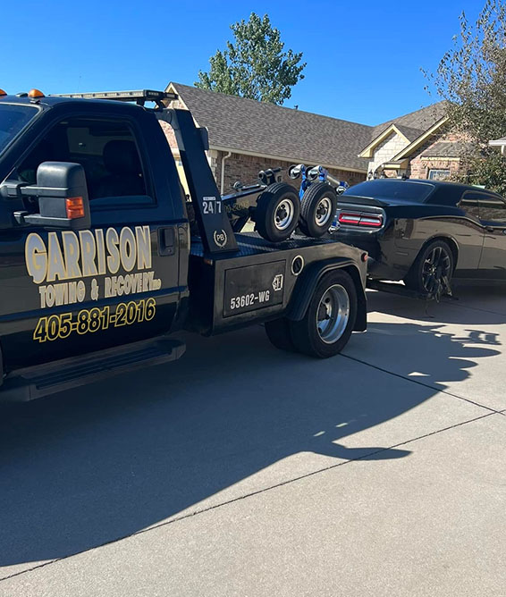 Tow truck from Garrison Towing & Recovery towing a black muscle car in a residential driveway on a sunny day.
