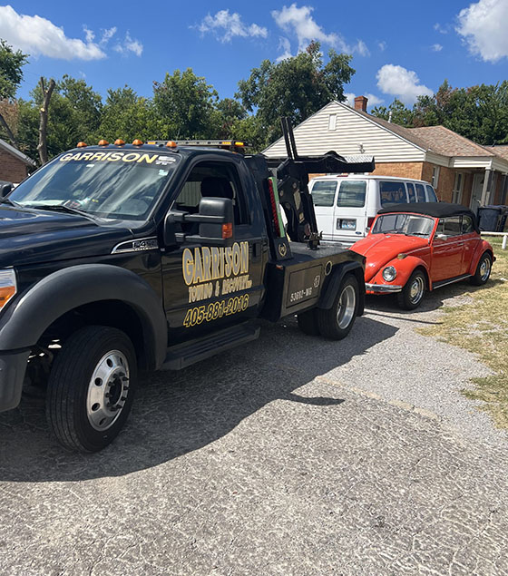 A black tow truck from Garrison Towing is towing a red vintage Volkswagen Beetle convertible on a suburban street.