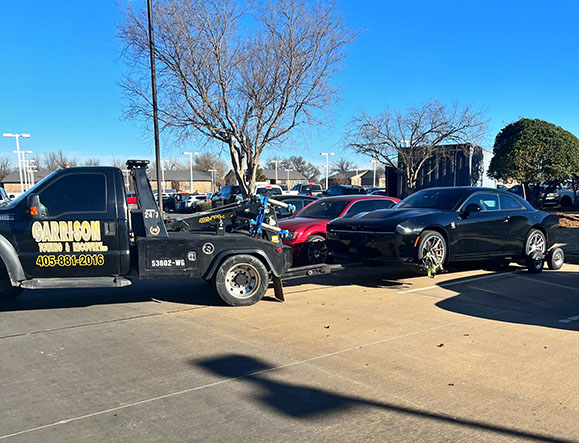 Tow truck from Garrison Towing & Recovery hauling a black sports car in a parking lot on a clear day.