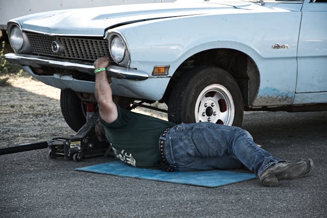 Person lying on a mat under a white vintage car, working on its front with a car jack supporting the vehicle.