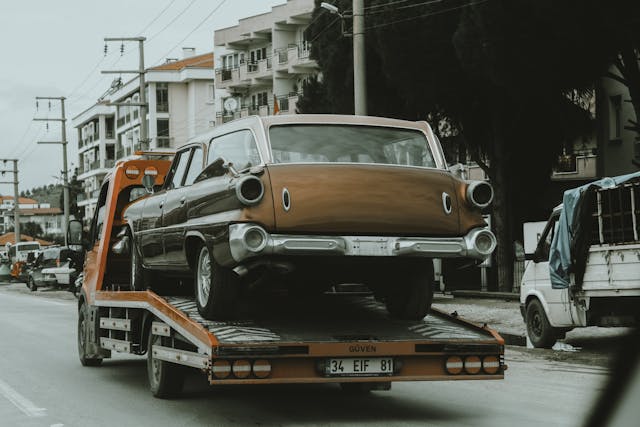 A vintage brown car being transported on a flatbed tow truck along a city street with buildings and other vehicles nearby.