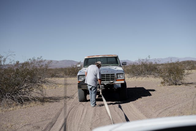 A man attaching a tow rope to a stuck white truck on a dry, desert trail with sparse bushes and distant mountains.