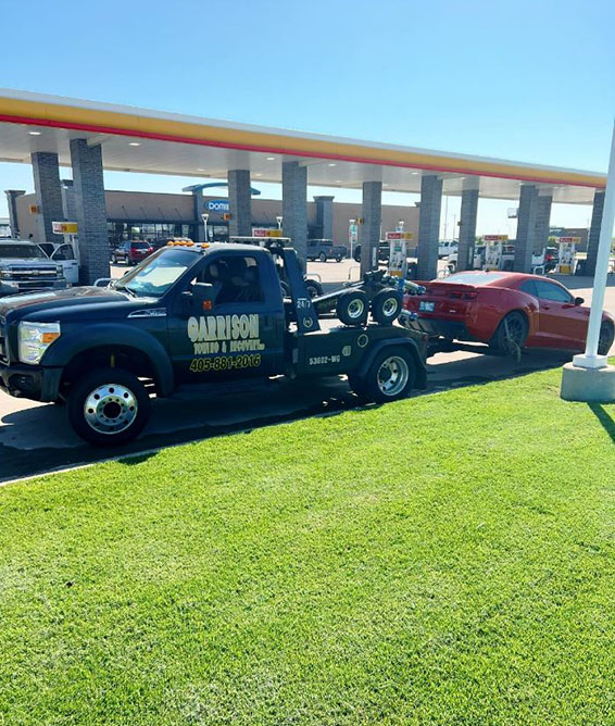 Black Garrison tow truck towing a red sports car at a gas station on a sunny day with green grass in the foreground.