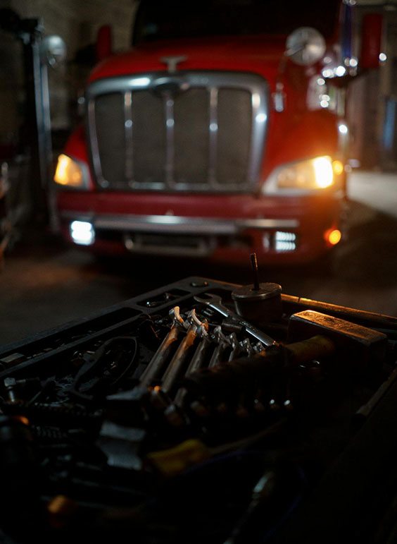 Red truck with headlights on in a dimly lit garage, foreground filled with various wrenches and tools on a workbench.